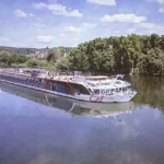 AmaRudi, Europe's largest river cruise ship, cruising along the Danube River with its modern double-width design, spacious sun deck, and scenic riverside landscape under a clear blue sky.