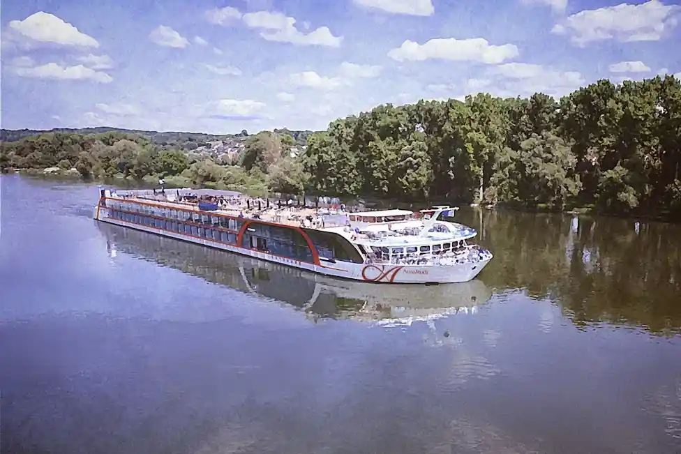 AmaRudi, Europe's largest river cruise ship, cruising along the Danube River with its modern double-width design, spacious sun deck, and scenic riverside landscape under a clear blue sky.