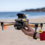A Surf Life Saving NSW operator holds a shark-spotting drone on a beach as Australia expands year-round drone patrols across 70 NSW beaches under a $34 million shark safety program with AI detection trials