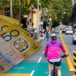 Cyclist riding in a Queensland bike lane with Australian $50 notes, illustrating Queensland's new e-bike laws, $518 fines, and road rule changes effective July 1, 2026.