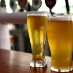Two glasses of beer sit on a wooden pub bar counter with beer taps blurred in the background, representing Queensland pubs preparing for extended trading hours during the Socceroos' FIFA World Cup match against Egypt