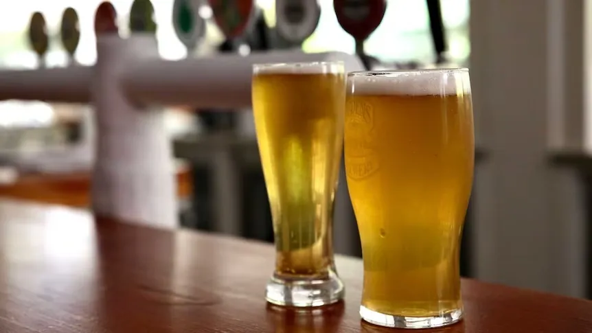 Two glasses of beer sit on a wooden pub bar counter with beer taps blurred in the background, representing Queensland pubs preparing for extended trading hours during the Socceroos' FIFA World Cup match against Egypt