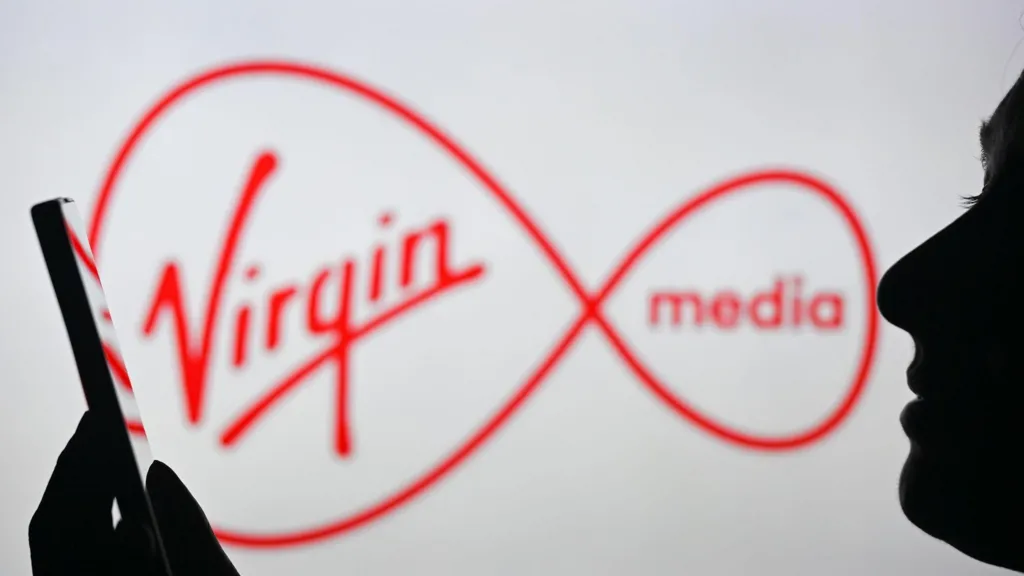 Virgin Media logo displayed on a screen behind a person using a smartphone, illustrating Ofcom's ÂŁ28 million fine over customer contract cancellation practices.