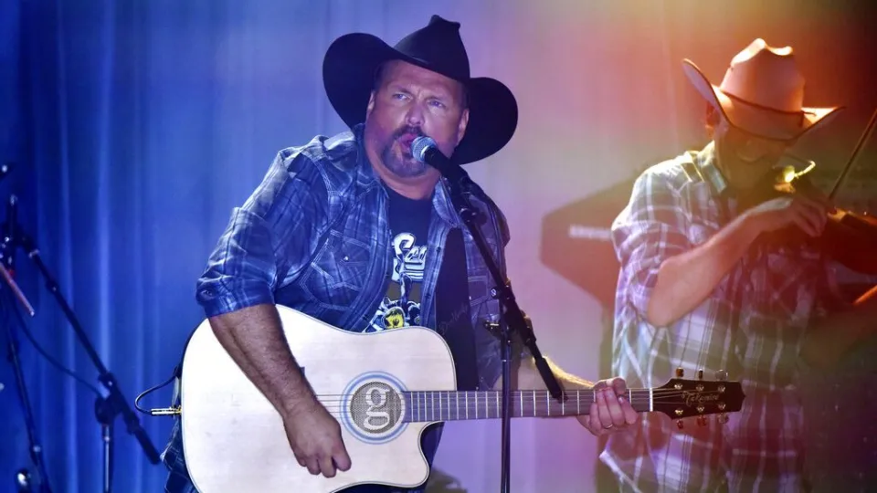 Garth Brooks performs on stage with an acoustic guitar during a live country music concert as a band member plays fiddle in the background.