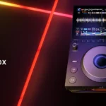 AlphaTheta CDJ-1500X compact DJ media player featuring a 10.1-inch touchscreen, illuminated jog wheel, and professional performance controls against a colorful studio lighting background.