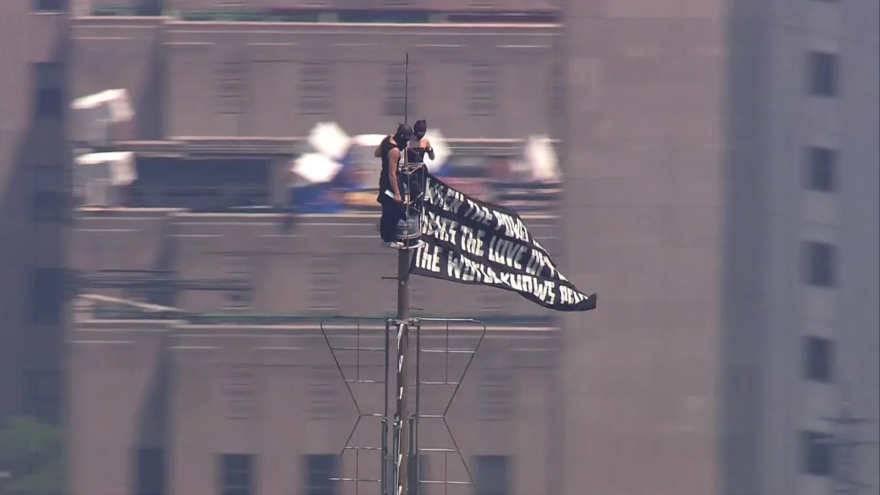 Two masked climbers stand atop the Empire State Building spire while unfurling a large peace banner high above Manhattan during an NYPD response in New York City on July 1, 2026.