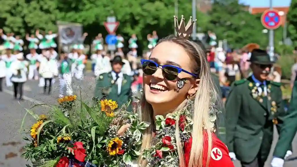 Young woman wearing a ceremonial crown smiles while holding flowers during the Hannover SchĂĽtzenfest Parade 2026 as marching bands and participants walk through Hanover city centre.