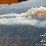 Aerial view of the fast-moving Los Gallardos wildfire in Almería, Spain, with thick smoke billowing over hills as firefighting aircraft monitor the blaze.