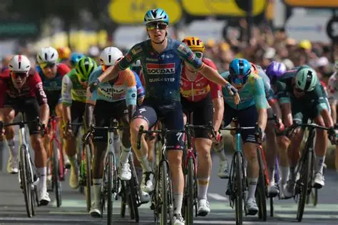 Tim Merlier celebrates winning Stage 8 of the 2026 Tour de France in Bergerac as the peloton crosses the finish line behind him.