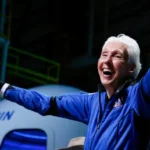 Wally Funk smiles with arms outstretched while wearing a blue Blue Origin flight suit in front of a New Shepard spacecraft after her historic space mission.