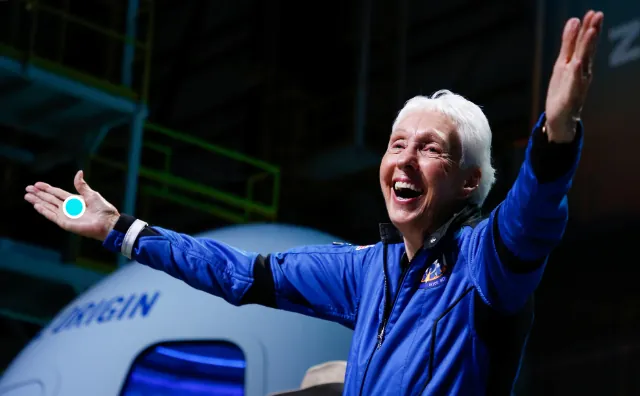 Wally Funk smiles with arms outstretched while wearing a blue Blue Origin flight suit in front of a New Shepard spacecraft after her historic space mission.